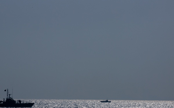 The image shows three vessels silhouetted against a sunlit open sea under a hazy gray sky. The largest vessel is positioned on the left, with two smaller vessels visible in the middle distance and far right. The strong backlighting creates a high-contrast silhouette effect with glittering light reflecting off the water surface.