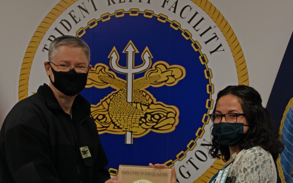 Two masked individuals are posing together while jointly holding an 'Employee of the Quarter' plaque at the Trident Refit Facility in Bangor, Washington. The person on the left appears to be in military/naval attire presenting the award to the person on the right. The ceremony takes place in front of the official Trident Refit Facility seal and flanking flags.
