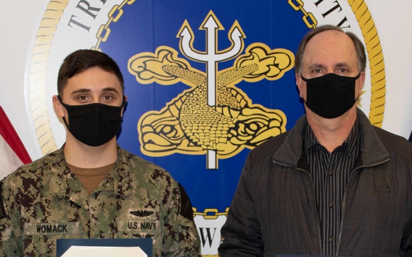 Two individuals stand before a U.S. Navy installation seal and an American flag, both wearing black face masks and holding official documents. The person on the left is a U.S. Navy sailor in NWU Type III camouflage uniform with the name tape 'WOMACK,' while the person on the right is a civilian holding a 'Special Achievement Award' certificate issued to 'John F. Walker' from Trident Base Facility, Bangor. This appears to be a formal military award ceremony.