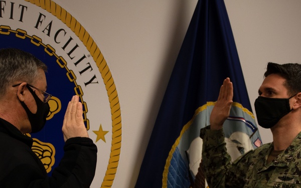 Two individuals are participating in what appears to be a military oath or swearing-in ceremony at a U.S. Navy Refit Facility. Both have their right hands raised, with one person in U.S. Navy digital camouflage (NWU Type III) uniform and the other in civilian black attire. Both are wearing black face masks, suggesting the photo was taken during the COVID-19 pandemic period.