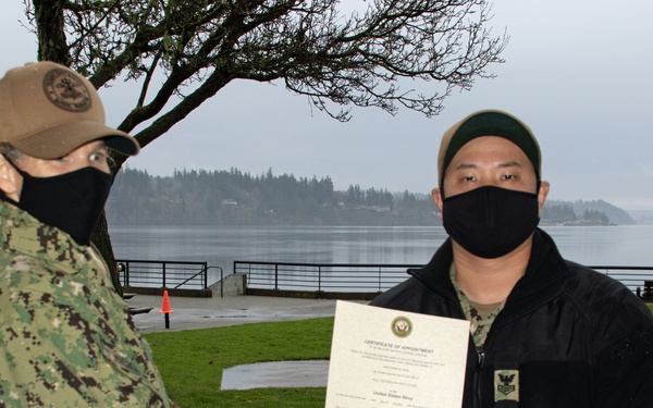 Two U.S. Navy personnel are shown outdoors near a waterfront, with one individual presenting a Certificate of Appointment to the other. Both individuals are wearing black face masks and military uniforms, suggesting this ceremony took place during the COVID-19 pandemic period. The recipient is holding the official document while shaking hands with the presenter.