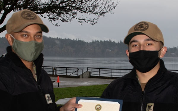 Two individuals wearing matching tan/khaki caps and black face masks are holding a Certificate of Appointment from the United States Navy. They are standing outdoors near a waterfront area with a railing, trees, and a body of water visible in the background. Both are dressed in black jackets and appear to have Navy insignia patches on their clothing.