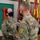 Two U.S. Army soldiers in OCP (Operational Camouflage Pattern) uniforms are shaking hands during what appears to be a military awards ceremony. The female soldier, identified by her name tape as 'SMITH,' is wearing a medal on her chest, suggesting she is receiving a commendation or award. Both individuals are wearing black face masks, indicating this event took place during the COVID-19 pandemic period.