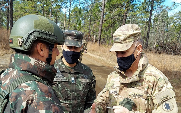 Three military personnel are engaged in a discussion on a dirt path in a pine forest setting. Two individuals wear Brazilian Army camouflage uniforms (one with a tactical helmet), while the third wears U.S. Army OCP (Operational Camouflage Pattern) uniform with visible 'U.S. ARMY' and 'RANGER' tab patches. All three are wearing dark face masks, suggesting this was taken during the COVID-19 pandemic period.