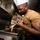 USS Ohio Chef Bakes a Treat A cook wearing a white chef's hat, black face mask, and tan t-shirt is pouring batter from a large stainless steel mixing bowl onto a sheet pan in what appears to be a commercial or military galley kitchen. The individual has visible tattoos on both arms. The setting suggests a professional kitchen environment, possibly aboard a naval vessel given the compact, utilitarian design.