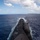 USS Ohio Operations Underway A large military submarine is photographed from an elevated aft position while underway on the surface of the ocean. The image shows the full length of the submarine's hull from stern to bow, with the sail (conning tower) visible midship and a coastline visible in the background. The vessel is creating white wake on both sides as it moves forward through deep blue open water.
