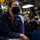 USS Ohio Operations Underway A U.S. Navy sailor wearing a black face mask, clear-framed glasses, and dark navy coveralls is seated in what appears to be a shipboard interior compartment. She displays a gold chief petty officer anchor insignia on her collar, indicating senior enlisted rank. Several other masked sailors in similar uniforms are visible in the background of the cramped, equipment-filled space.