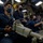 USS Ohio Operations Underway Military personnel in dark blue coveralls are stationed at control consoles inside what appears to be a submarine control room. All visible crew members are wearing black face masks, suggesting this was taken during the COVID-19 pandemic period. The crew members are seated at various control stations with complex instrumentation panels and control mechanisms visible throughout.