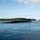 USS Ohio Operations Underway A large military submarine is seen traveling on the surface of calm blue waters near a coastline. The submarine appears dark/black in color with a visible sail (conning tower) and antenna masts. A green, tree-covered landmass and a smaller rocky outcrop are visible in the background under a partly cloudy sky.