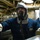 USS Ohio Operations Underway A person wearing a full-face respirator/gas mask, gray hood, blue coveralls, and white gloves is working in what appears to be a confined mechanical or engineering space. A name tag and qualification badge are visible on the coveralls. A second similarly equipped individual is partially visible in the background to the right.