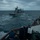 USS BARRY and JMSDF JS TOWADA Night Replenishment at Sea A naval replenishment-at-sea (RAS) or approach operation is underway between two warships on open, overcast seas. Personnel on the foreground vessel's bridge wing are monitoring and coordinating the approach of a larger auxiliary or supply-type vessel ahead. Crew members in dark naval uniforms are visible on the bridge wing, with one holding a whiteboard and another operating equipment.
