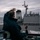 USS BARRY and JMSDF JS TOWADA Night Replenishment at Sea Two naval personnel are visible from behind on the bridge wing of a warship, with one individual raising their hand in a wave or signal gesture. A large gray naval vessel is positioned nearby in open sea waters under overcast skies. The person in the foreground wears a dark jacket with text that appears to read 'DDG52 CO', suggesting this may be the Commanding Officer of USS Barry (DDG-52).