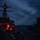 USS BARRY and JMSDF JS TOWADA Night Replenishment at Sea A naval warship, appearing to be a U.S. Navy Arleigh Burke-class guided-missile destroyer, is conducting an underway replenishment (UNREP) operation with a supply vessel at dusk or dawn. Fuel or supply lines are visibly strung between the two vessels as crew members work on deck under low-light conditions. The scene is dramatically lit by red operational lighting and ambient twilight against an overcast sky.