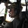 75th EAS moves cargo in East Africa A military aviator is seated in an aircraft cockpit, wearing a multicam/OCP pattern cap, Bose aviation headset, sunglasses, and tactical gear. The cockpit displays green avionics screens including what appears to be a radar display and navigation instruments. The pilot appears to be in flight, looking forward through the cockpit windscreen.