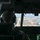 75th EAS moves cargo in East Africa The image shows the interior of a military aircraft cockpit during flight, with at least one crew member visible from behind wearing a flight helmet and headset. Through the windscreen, a coastal runway and urban area are visible below, suggesting an approach or departure from an airfield near the ocean. The cockpit features multiple green-lit digital displays and an extensive overhead panel with switches and controls.
