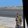75th EAS moves cargo in East Africa A military service member in OCP (Operational Camouflage Pattern) uniform stands on an aircraft ramp/taxiway area at what appears to be a military airfield. The individual is wearing a communications headset, holding a blue binder/checklist, and managing interphone cables, suggesting they are performing pre-flight or maintenance ground crew duties. The arid, sparse landscape and runway signage are visible in the background.