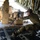 75th EAS moves cargo in East Africa A military service member in MultiCam/OCP camouflage uniform is securing cargo chains inside what appears to be a C-130 Hercules military transport aircraft. The individual is physically straining to fasten or tighten heavy-duty chains attached to a large piece of tan/desert-colored military equipment being loaded or secured. A second service member wearing sunglasses and a headset is partially visible in the background.