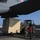 75th EAS moves cargo in East Africa Military or government cargo loading operation at an airfield, showing ground crew directing a front-end loader carrying a large tan/beige cargo container toward a large military transport aircraft. Two ground personnel are guiding the forklift operator as the container is positioned beneath the aircraft's wing. The setting appears to be a warm, arid location based on the clear blue sky and building architecture visible in the background.