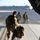 75th EAS moves cargo in East Africa Three military personnel in multicam/OCP camouflage uniforms are disembarking from what appears to be a military transport aircraft via a rear cargo ramp onto a tarmac. Two of the soldiers are wearing flight/communications headsets, and one is carrying a rifle. A civilian ground crew member in a high-visibility vest is visible in the background on the airfield.