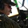 75th EAS moves cargo in East Africa A military aviator is seated in a helicopter cockpit, wearing multicam/OCP camouflage uniform with a US flag patch visible on the shoulder. He is equipped with Bose aviation headset, a boom microphone, and tinted aviator sunglasses, suggesting active flight operations. The cockpit displays green-lit instrumentation panels visible in the background.
