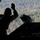 A silhouetted military crew member is positioned at the open rear ramp of what appears to be a military helicopter or transport aircraft in flight. The individual has one hand raised, likely signaling during an aerial operation such as a cargo drop or airdrop. A forested landscape is visible below through the open ramp.