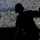 A silhouetted figure wearing a helmet, likely a military jumpmaster or aircrew member, is visible in the foreground against a backdrop of dense forest. In the background, at least one open parachute canopy is visible descending toward the tree line, indicating an active airborne/parachute operation. The image is taken from inside an aircraft, likely during a military jump exercise.