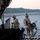 Two military personnel in multicam/OCP camouflage uniforms are standing on or near the rear cargo ramp of a large military transport aircraft, likely a C-130 or similar platform, on an airfield at dusk. One individual faces the camera wearing a flight helmet with night vision device mount, while the other faces away carrying a tactical backpack and what appears to be a coiled hose or equipment bundle. The setting appears to be a forward operating or training airfield with a treeline visible in the background.