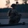 A military service member in full tactical gear is kneeling on what appears to be a tarmac or airfield surface during dusk or dawn. The soldier is equipped with a combat helmet mounted with night vision device, multicam camouflage uniform, body armor, and a backpack. A large hose or fuel line is visible on the ground nearby, suggesting possible aircraft refueling or airfield operations.