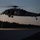 A U.S. Navy helicopter, identifiable by 'NAVY' markings on its fuselage, is captured hovering at low altitude over a tarmac or runway during dusk or dawn conditions. The scene is dramatically lit by the helicopter's own searchlight reflecting off the ground surface. A second aircraft is faintly visible in the background to the right.