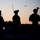 The image shows silhouettes of approximately four military personnel standing at dusk or dawn against an orange-pink gradient sky. The soldiers are equipped with helmets mounted with night-vision devices and radio antennas, suggesting a special operations or tactical unit. Two aircraft are visible in the background sky, and small indicator lights (white and green) are visible on the personnel.