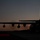 A large military turboprop aircraft is silhouetted against a twilight sky on a tarmac or airfield. The image is taken during dusk or dawn, with a warm orange-red glow near the horizon and a deep blue-gray upper sky. The aircraft's rear cargo ramp appears to be open, and ground equipment is visible nearby.