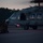 A military helicopter, appearing to be an HH-60 Pave Hawk or similar variant, is on a tarmac at dusk or dawn with rotor blades spinning. Two armed and helmeted personnel in tactical gear are kneeling on the ground in front of the aircraft, appearing to be preparing for or concluding an operation. The low-light conditions and active rotor suggest an imminent insertion or extraction scenario.