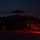 A military helicopter, consistent with a Sikorsky UH-60 Black Hawk or similar variant, is photographed on a tarmac or landing zone at night. Two personnel in tactical gear are standing near the aircraft, which has its rotors spinning as indicated by motion blur. The scene is illuminated primarily by red and green navigation/warning lights, creating a low-light operational atmosphere.