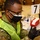 Michigan’s Task Force Red Lion, Administers the COVID-19 Vaccine A military service member in camouflage uniform and high-visibility vest is administering an injection to a patient at what appears to be a mass vaccination or medical event. The individual is wearing black nitrile gloves, a black face mask, and glasses while carefully inserting a syringe into a patient's arm. A numbered station marker '7' is visible in the background along with medical supplies on a table.