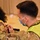 Michigan’s Task Force Red Lion, Administers the COVID-19 Vaccine A person in military camouflage uniform and a high-visibility yellow vest is administering an injection into the upper arm of a civilian woman wearing a yellow-green cardigan and a black face mask. Both individuals are wearing face masks, suggesting this is a medical or vaccination event. The setting appears to be an indoor venue, likely a mass vaccination site.