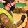 Michigan’s Task Force Red Lion, Administers the COVID-19 Vaccine A U.S. Army soldier in OCP (Operational Camouflage Pattern) uniform and high-visibility vest is administering a vaccine injection to a civilian recipient. The setting appears to be a mass vaccination site, likely during the COVID-19 pandemic based on the face masks worn by both individuals. The soldier is wearing black nitrile gloves and using a syringe to inject the recipient's upper arm.