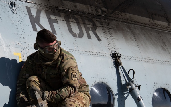 A military service member in full camouflage uniform and protective eyewear is crouched on top of a helicopter, performing what appears to be a refueling operation. The aircraft is marked with 'KFOR' (Kosovo Force), indicating a NATO peacekeeping mission context. The soldier is handling a fuel nozzle/hose near a fuel port labeled 'JET A-1'.