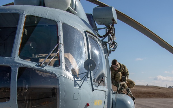 Military personnel are conducting a refueling operation on a light blue/gray helicopter on a tarmac or airfield. One person is actively handling a fuel hose on top of the helicopter while a second individual stands nearby observing. A red fire extinguisher is positioned on the ground as a safety precaution during the fueling process.