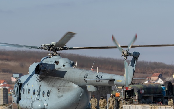 A large military helicopter, marked with the number '261' and a checkered emblem consistent with Croatian armed forces markings, is parked on a tarmac at a military airfield. Ground personnel in camouflage uniforms are present nearby, and a fuel tanker truck is visible in the background, suggesting a refueling operation is underway. The setting appears to be a forward operating base or military airfield in a hilly, semi-rural area.