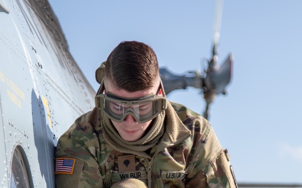 A U.S. Army soldier named Wilbur, wearing OCP camouflage uniform and protective goggles, is crouched on top of a military helicopter performing a refueling operation. The soldier is handling a fuel nozzle/coupling assembly, with text on the aircraft fuselage partially reading 'JET A' indicating jet fuel type. A helicopter rotor is visible in the background.