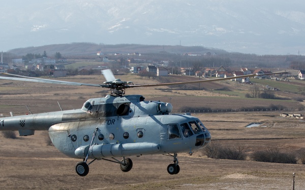 A light blue military helicopter marked '251' with a Croatian checkerboard insignia and 'KFOR' markings is captured hovering just above a tarmac surface, likely during takeoff or landing. The aircraft appears to be a Mil Mi-8/Mi-17 type helicopter operating under NATO's Kosovo Force (KFOR) mission. The background shows a rural landscape with scattered buildings, bare trees, and snow-capped mountains in the distance.