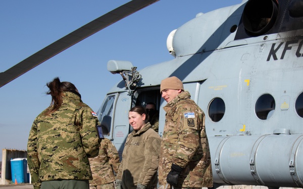 Military personnel in various camouflage uniforms are gathered on a tarmac next to a light gray helicopter marked 'KFOR', indicating a NATO Kosovo Force operation. The group appears to be engaged in conversation or a briefing near the aircraft. The setting is an outdoor airfield under clear blue skies in cold weather conditions.