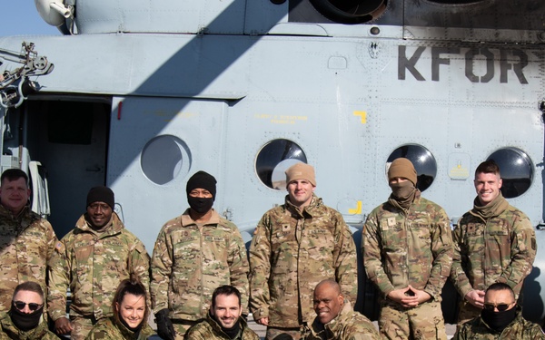 A group of approximately eleven military personnel in camouflage uniforms pose for a group photo in front of a large helicopter marked with 'KFOR' (Kosovo Force - NATO-led peacekeeping mission). The personnel are arranged in two rows, with six standing in the back and five kneeling in the front, on what appears to be a military airfield or tarmac. The helicopter behind them is a large transport/utility aircraft painted in light gray.