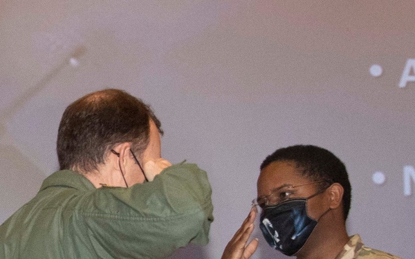 Two military personnel are exchanging salutes during what appears to be an award ceremony. The individual on the right, wearing a US Air Force OCP (Operational Camouflage Pattern) uniform, is holding a decorative eagle and American flag trophy/award. Both individuals are wearing face masks, suggesting this event took place during the COVID-19 pandemic period.