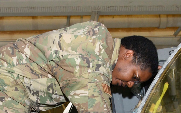 A military service member wearing OCP (Operational Camouflage Pattern) uniform is leaning over and inspecting the cockpit area of a military aircraft. The individual appears to be conducting a maintenance check or pre-flight inspection inside a hangar facility. A US flag patch is visible on the right sleeve of the uniform.