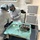 A medical or dental surgical procedure is being performed by two healthcare professionals wearing sterile attire including surgical caps, masks, gloves, and white coats. A sterile instrument tray covered with a green surgical drape is prominently visible in the foreground, laid out with various surgical tools. The setting appears to be a clinical or surgical room equipped with medical storage units and air filtration equipment.