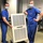 Two healthcare workers in blue scrubs and surgical masks pose with a large white air purification unit on a wheeled stand in what appears to be a medical or clinical facility. Both individuals are smiling beneath their masks and appear to be presenting or showcasing the equipment. The setting includes office furniture, bulletin boards, and posted notices visible in the background.