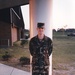 A young male service member stands at attention in front of a white column on a concrete walkway, wearing woodland camouflage BDU (Battle Dress Uniform) and black combat boots. The setting appears to be a military installation or training facility, with a brick barracks-style building visible to the left and an open grassy field to the right. The photo has the warm tones and grain characteristic of consumer film photography from the 1990s.