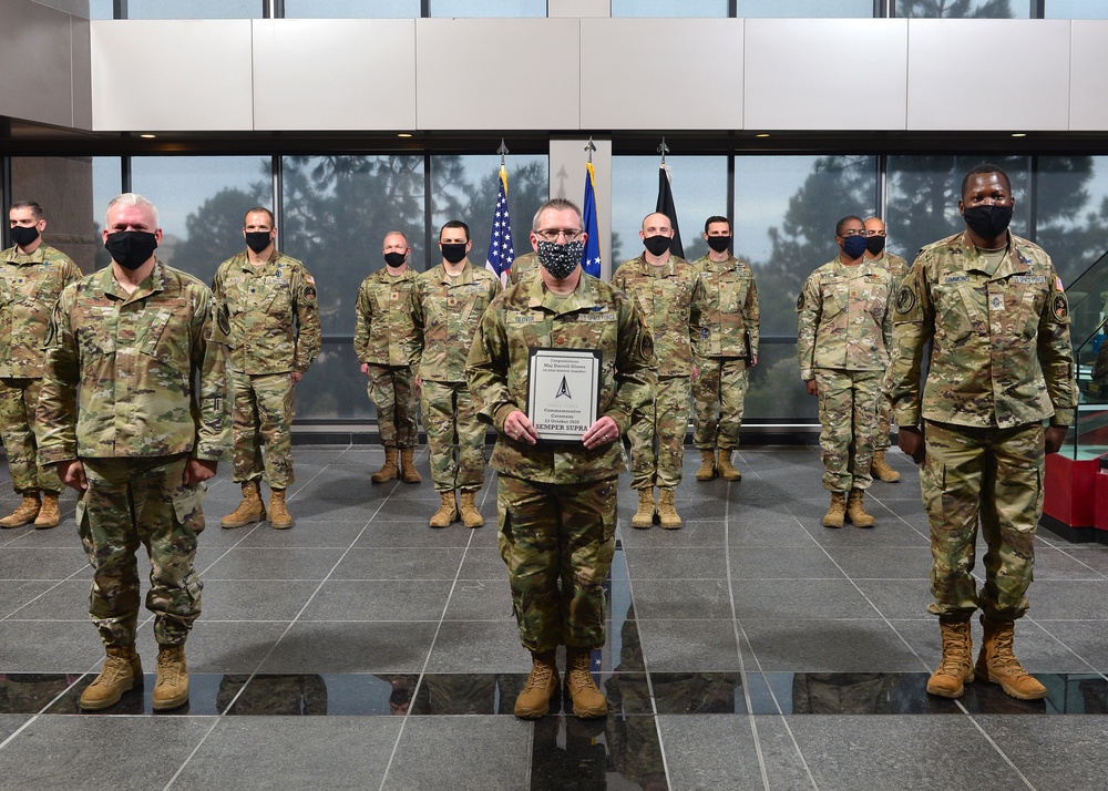 A group of approximately 12 military personnel in OCP (Operational Camouflage Pattern) uniforms are posed for a formal group photo inside a modern building. The central figure holds a commemorative plaque bearing the text 'SEMPER SUPRA' and the Space Force delta insignia. All personnel are wearing black face masks, suggesting this photo was taken during the COVID-19 pandemic period.
