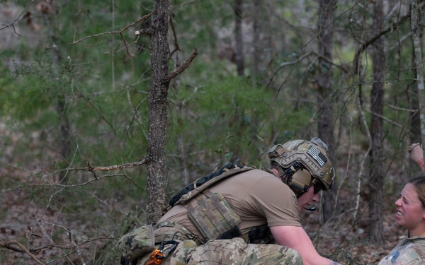Two individuals in military/tactical camouflage uniforms are positioned at the base of a large tree in a wooded area. One person wearing full tactical gear including a helmet, plate carrier, and communications equipment is crouching over a second person who is seated against the tree and appears to be in distress or playing a casualty role. The scene is consistent with a military combat casualty care training exercise.